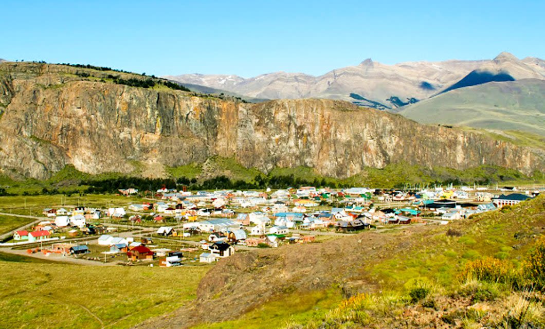 A small town with colorful houses is nestled in a green valley near El Chalten, surrounded by rocky cliffs and distant mountains under a bright blue sky—perfect for joining a trekking tour.