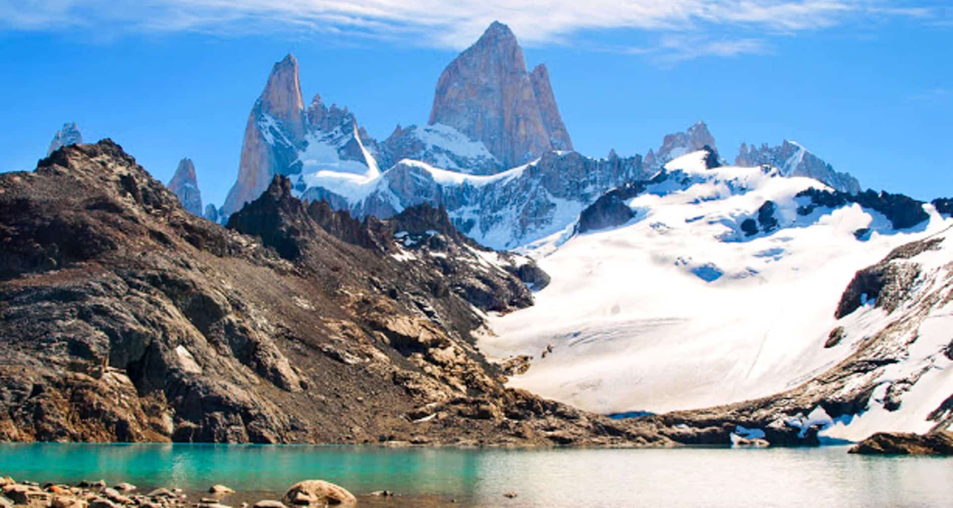 Snow-capped jagged mountains rise behind a rocky foreground and a turquoise lake near El Chalten, under a bright blue sky with scattered clouds—a breathtaking view on any Patagonia Eco Tour.