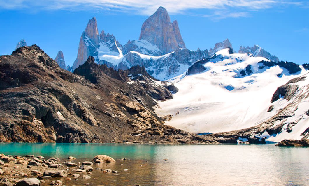 Snow-covered mountains with jagged peaks rise above turquoise water and rocky terrain under a blue sky with wispy clouds—an iconic scene from a Trekking Tour between El Calafate and El Chalten.