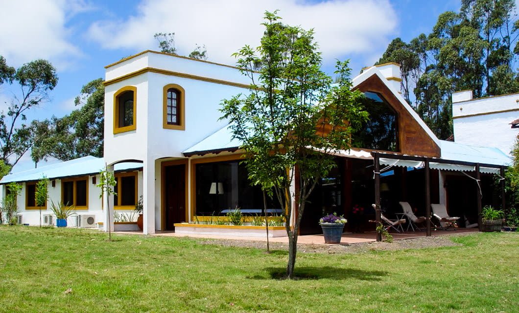 A large, modern house with white and yellow walls, blue roof, and many windows sits on a green lawn with trees and patio furniture, perfect for relaxing after a Punta del Este wine tour, under a clear, partly cloudy sky.