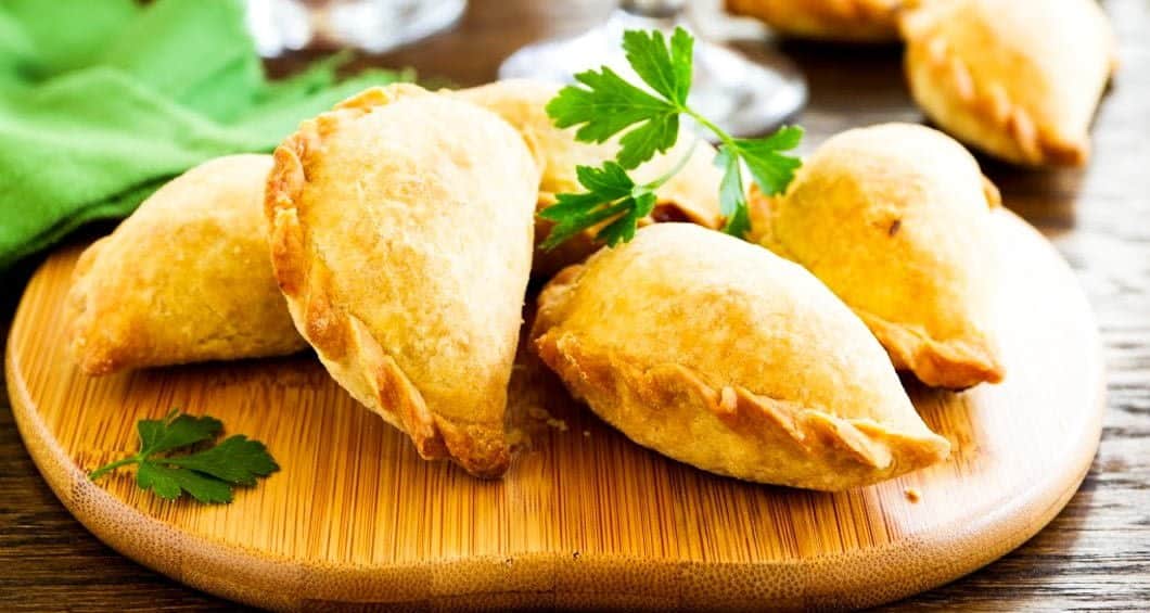 A wooden board with several golden-brown empanadas, garnished with fresh parsley, sits on a rustic wooden table with a green napkin in the background—perfect for savoring during your Argentina Chile travel adventure.