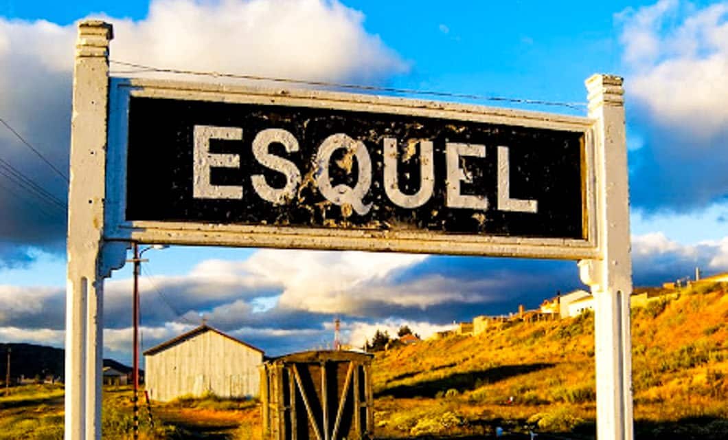 A weathered sign reading ESQUEL stands outdoors, with golden sunlight illuminating nearby small buildings and grassy hills—perfect for travelers heading to Bariloche or embarking on wildlife watching adventures under a partly cloudy sky.