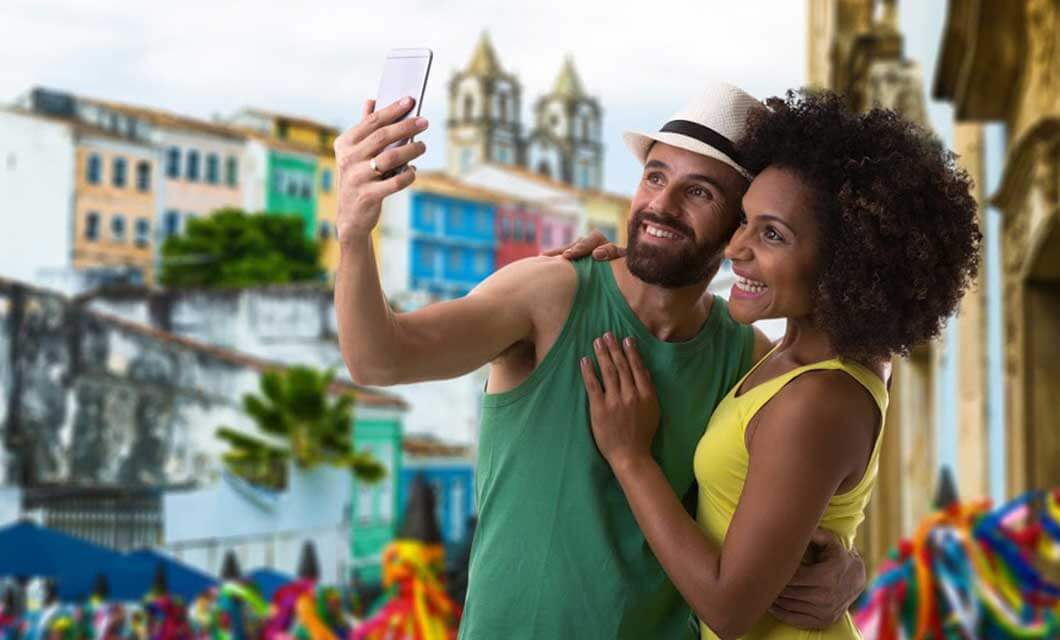 A smiling couple takes a selfie outdoors in front of colorful buildings and festive decorations, capturing the vibrant spirit of a Brazil's Cerrado tour—perhaps exploring Jalapão during a lively cultural or street festival.