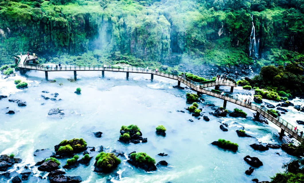A winding pedestrian bridge stretches over a misty river with scattered rocks and green vegetation, surrounded by lush forest and a waterfall in the background—an enchanting scene along the Brazil Gold Route.