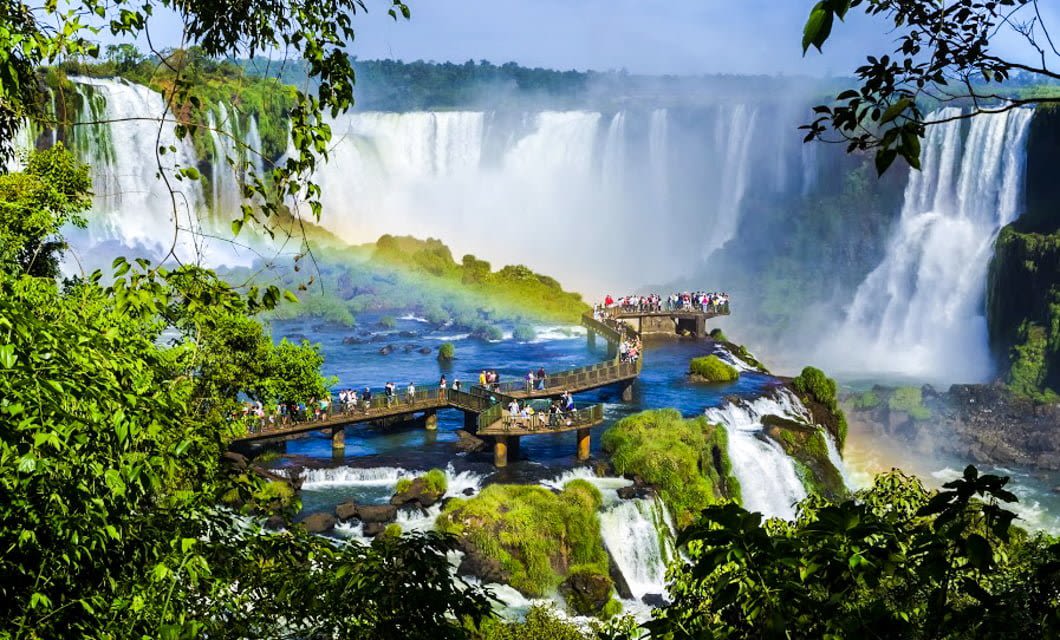 Tourists stand on a boardwalk in Argentina’s Land of Contrast, surrounded by lush greenery overlooking massive waterfalls and a rainbow at Iguazu Falls, with mist rising and forest in the background.