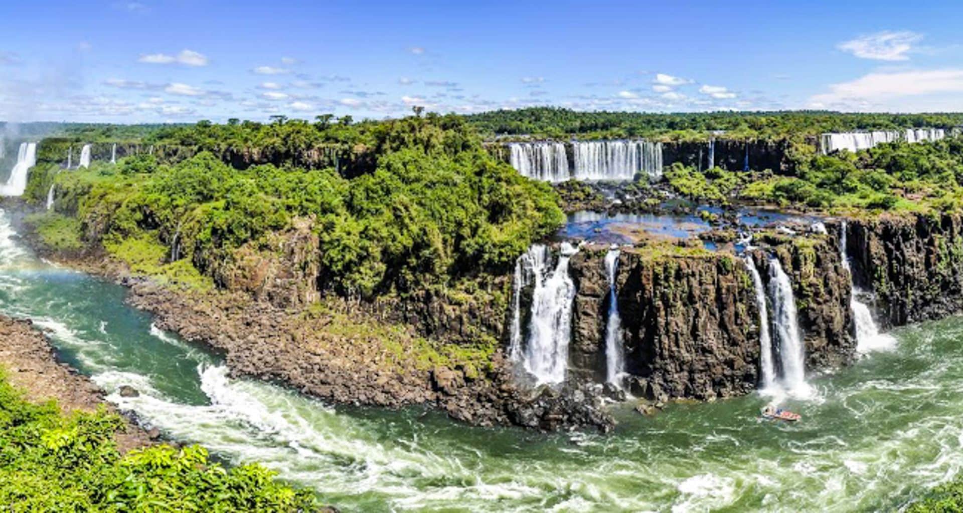 A panoramic view of Iguazu Falls, one of the World Wonders of South America, with multiple waterfalls cascading over lush, green cliffs into a winding river under a bright blue sky.