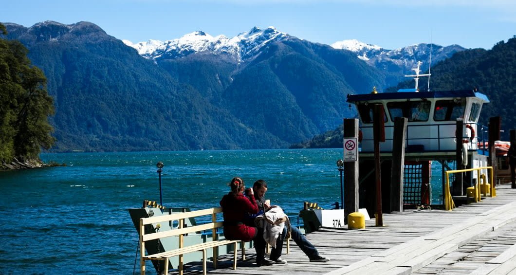 Two people sit on a bench on a wooden dock by a lake, with a boat moored nearby—reminiscent of the scenic Argentina to Chile Lake Crossing, surrounded by snow-capped mountains and lush forests under a clear blue sky.