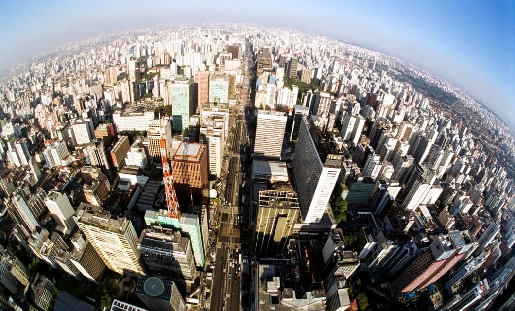 A wide-angle aerial view of a densely packed city with numerous tall buildings and skyscrapers under a clear blue sky, stretching out to the horizon—a striking contrast to the landscapes seen on a Brazil farm tour.