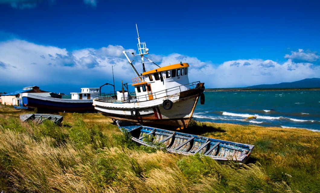 Three abandoned boats rest on grassy land near a windy shoreline in Chilean Patagonia, under a bright blue sky with waves on the water and distant hills in the background.