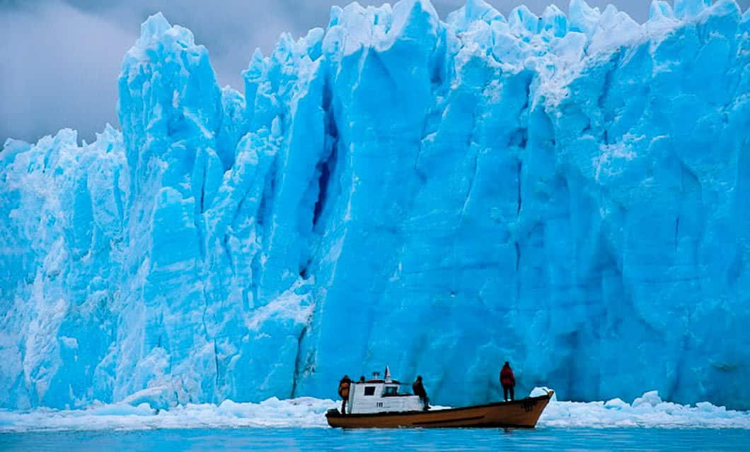 A small boat with people on board floats near a towering, jagged blue glacier in the Chilean Fjords, surrounded by icy water under a cloudy sky.