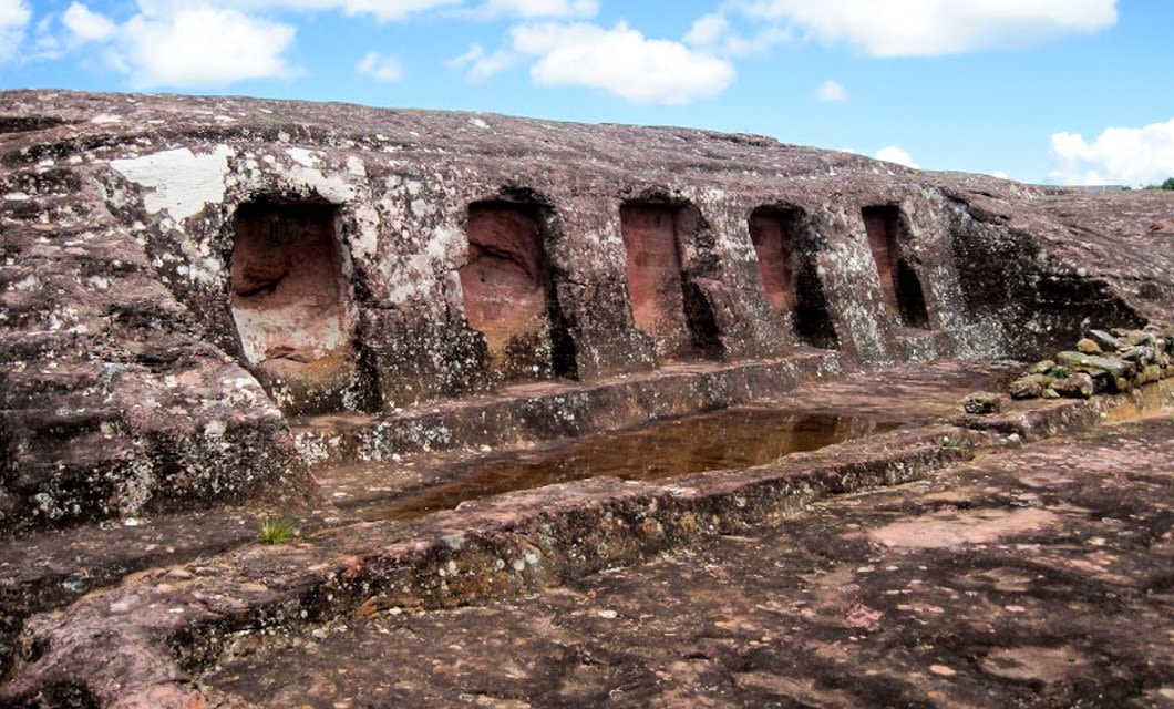 A rocky surface features six rectangular niches carved into the stone, with a shallow, rectangular pool of water in front—one of the Bolivia highlights to discover under a bright, partly cloudy sky on your Bolivia tour.