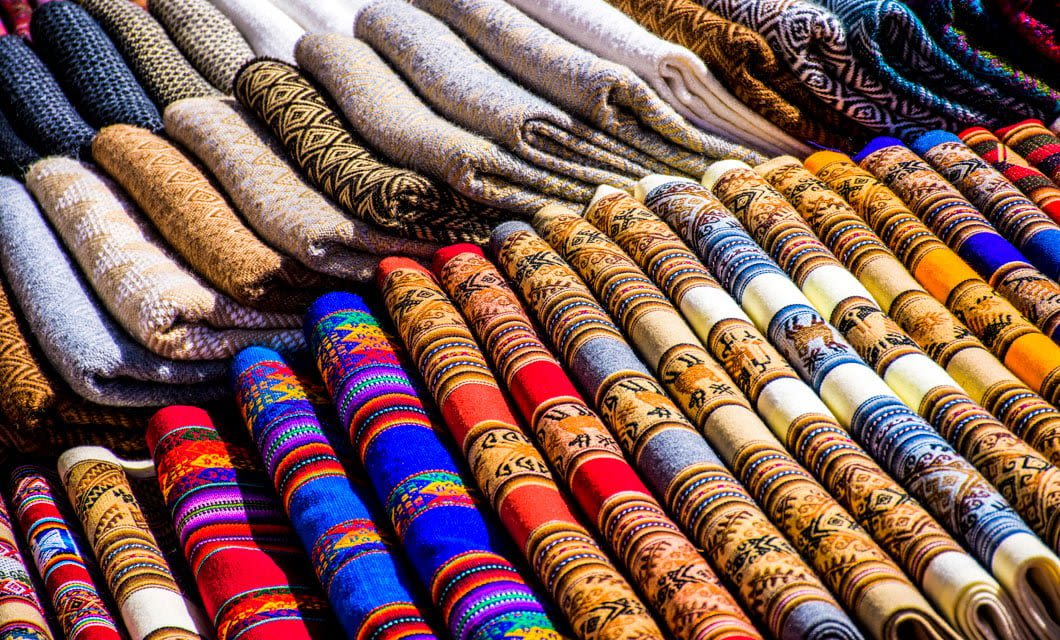 Colorful rows of folded traditional textiles, including shawls and blankets with intricate designs, displayed on a market stall—an authentic sight on a South America Tour.