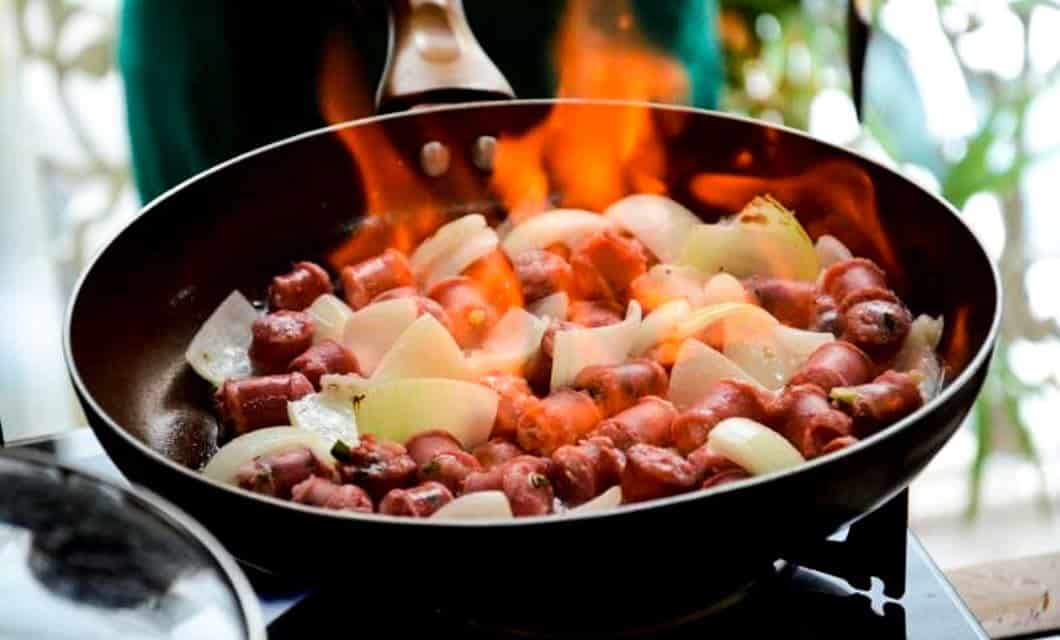 A close-up of sliced sausages and chopped onions sizzling in a frying pan with visible flames rising, capturing the bold flavors and fiery cooking style often found in Brazilian food.