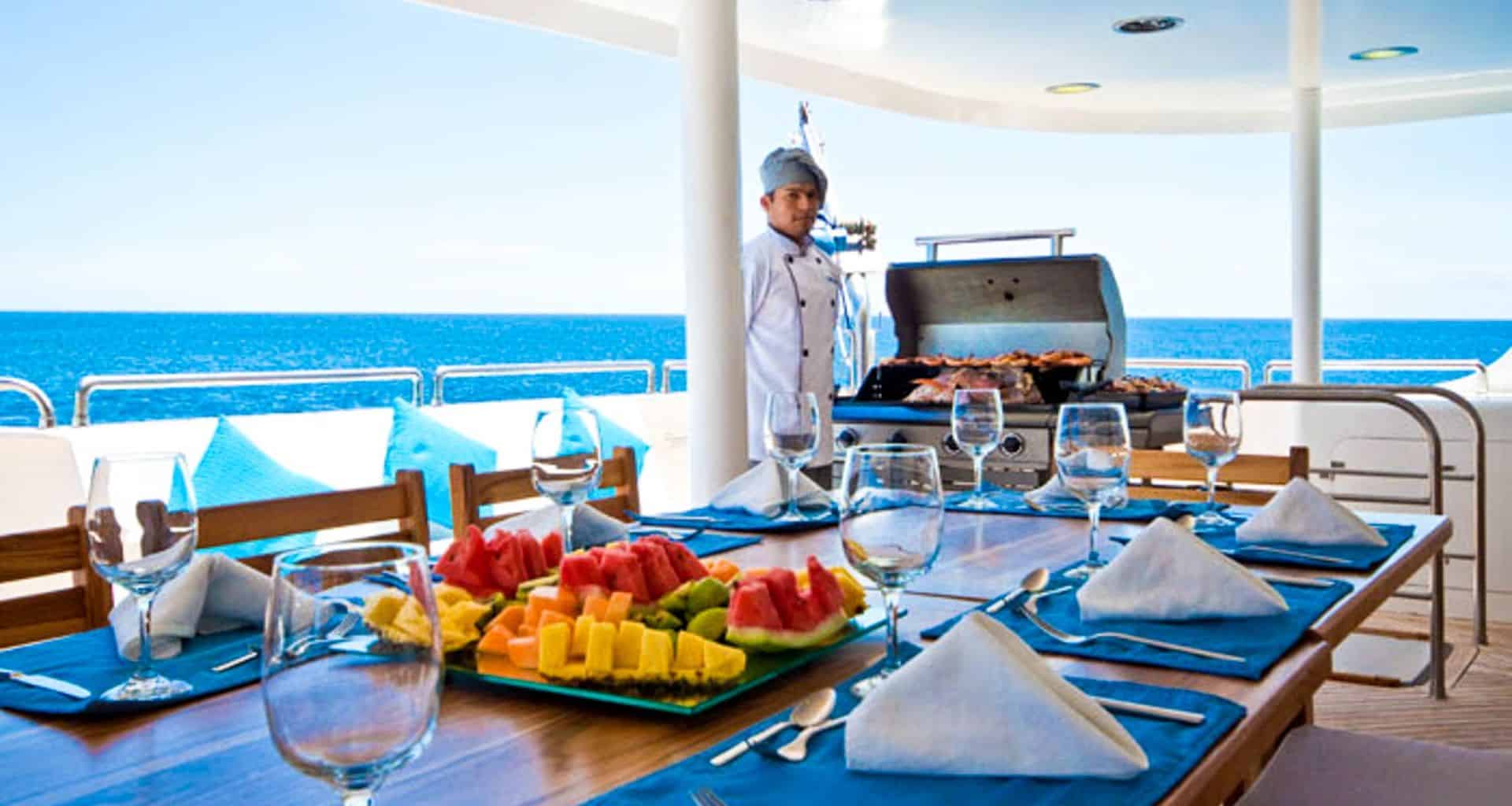 A chef grills food on the deck of the Cormorant Galapagos Cruise yacht by the ocean. A wooden table is set with wine glasses, napkins, and tropical fruits. Blue seat cushions and clear skies complete this idyllic scene.