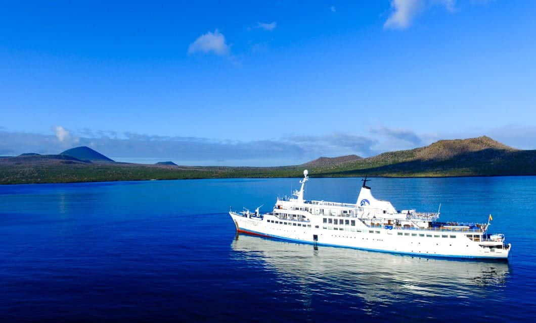 A white Galapagos Legend cruise ship sails on calm blue waters, framed by lush green hills and distant mountains under a bright blue sky with a few clouds.