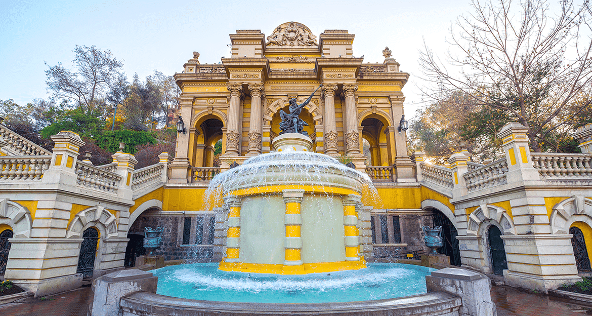 A grand yellow-and-white baroque-style building with ornate columns and arches stands behind a large tiered fountain, surrounded by staircases and sculptures—an architectural gem among the Wonders of Chile, with trees in the background.