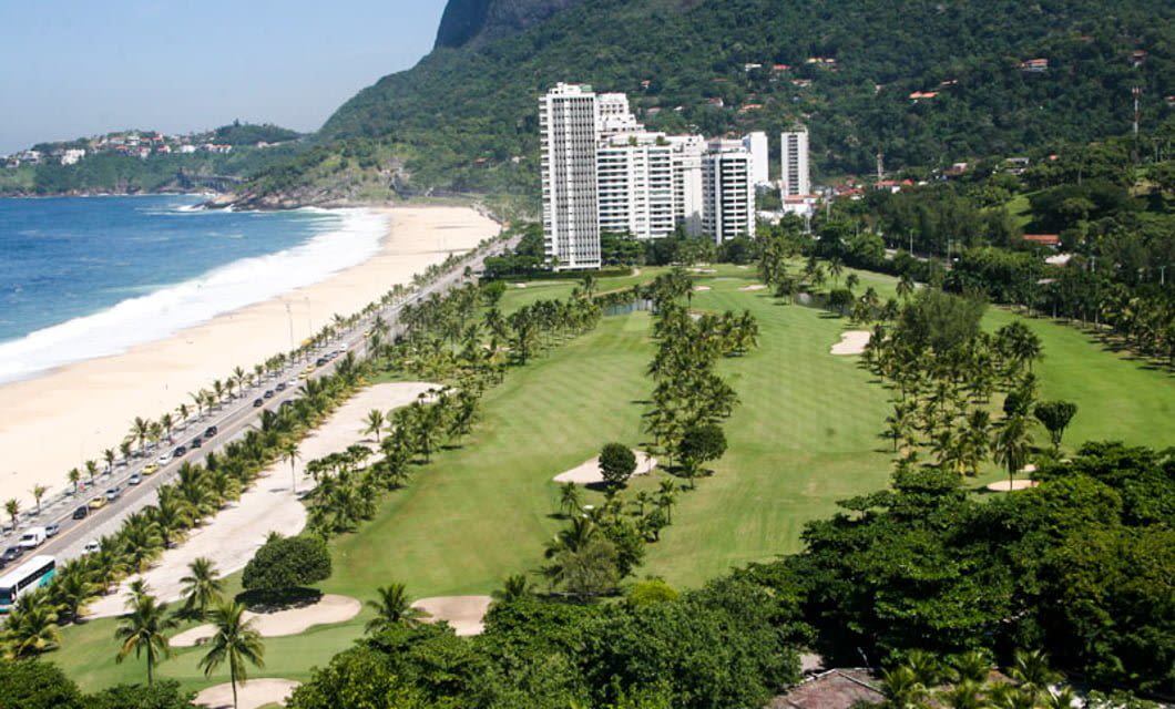 Aerial view of a coastal golf course, perfect for your next Brazil Golf Tour, with palm trees beside a sandy beach and ocean. High-rise buildings and green hills form the backdrop while a road runs parallel to the beach.