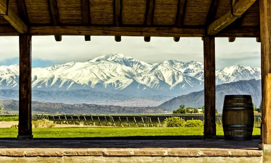 View of snow-capped mountains seen through a wooden porch with a barrel on the right side and green vineyards stretching out in the foreground, capturing the essence of Mendoza wine country perfect for wine tasting.