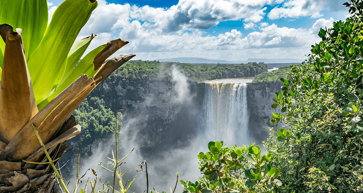 A wide waterfall cascades over a lush green cliff surrounded by forest, with mist rising above the falls—an ideal scene for a Three Guianas adventure tour. Large tropical leaves frame the view under a bright, partly cloudy sky.