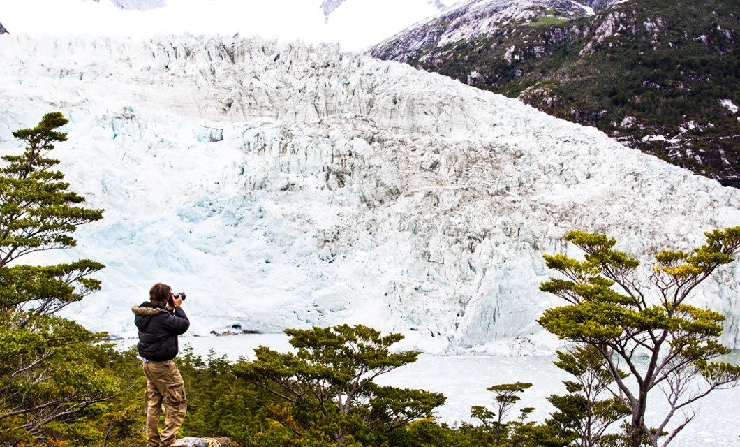 A person stands among green trees in Patagonia, photographing a massive, pale glacier with snowy mountains and a cloudy sky in the background.