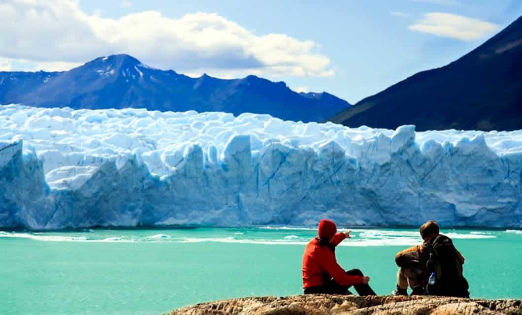 Two people in outdoor gear sit on a rocky surface, overlooking a turquoise lake and vast glacier in Chilean Patagonia, framed by snow-capped mountains under a partly cloudy sky.