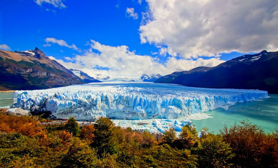 A massive glacier extends into turquoise water in Deluxe Patagonia, surrounded by rugged mountains and dense, colorful foliage under a partly cloudy blue sky.