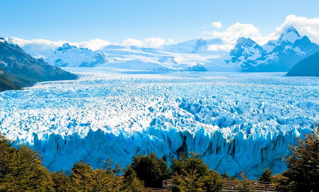 A vast, bright blue glacier stretches across the landscape in Patagonia, bordered by jagged snow-capped mountains under a clear blue sky, with green trees in the foreground.