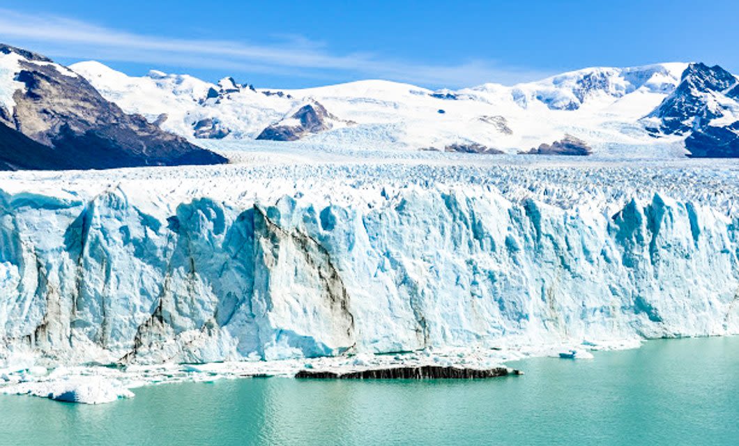 A vast glacier with jagged ice cliffs rises above turquoise water, backed by snow-covered mountains under a clear blue sky—an iconic scene near El Calafate on a Patagonia Eco Tour.