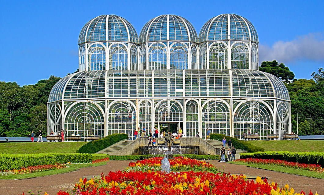 A large glass and metal greenhouse with three domes, surrounded by gardens and colorful flower beds, under a clear blue sky. Reminiscent of Brazil farm visits, several people are walking near the entrance and around the fountain.