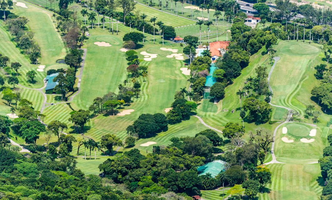 Aerial view of a golf course with several fairways, sand bunkers, greens, trees, and buildings surrounded by lush greenery and palm trees—an inviting paradise for golf courses lovers.