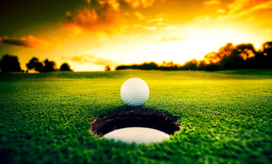 A golf ball rests at the edge of a hole on a green course during a vibrant sunset, capturing the serene beauty often experienced on a Brazil Golf Tour, with the sky glowing orange and yellow in the background.