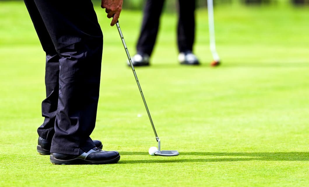 Close-up of a golfer’s legs and hands as they line up a putt on a green during a Golf Tour in Punta del Este, golf ball near the putter, with another golfer in the background, both wearing dark pants and shoes.