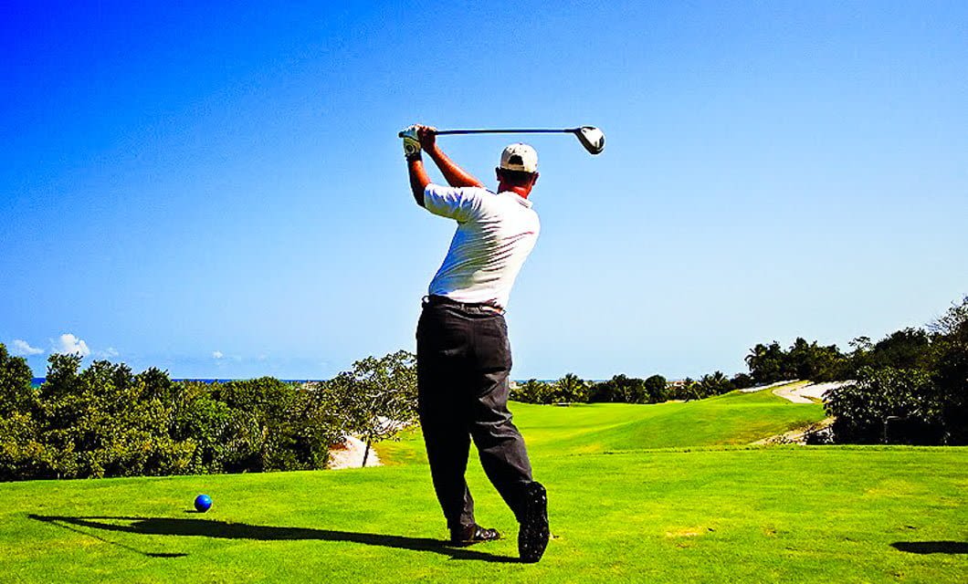 A golfer wearing a white shirt and cap swings a club on a bright green fairway under a clear blue sky, surrounded by trees and a distant sand trap during the South America Golf Tour.