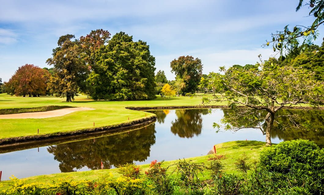 A scenic course perfect for a Brazil Golf Tour, with lush green fairways, sand bunkers, a curved water hazard reflecting trees, and vibrant landscaping under a blue sky with scattered clouds.