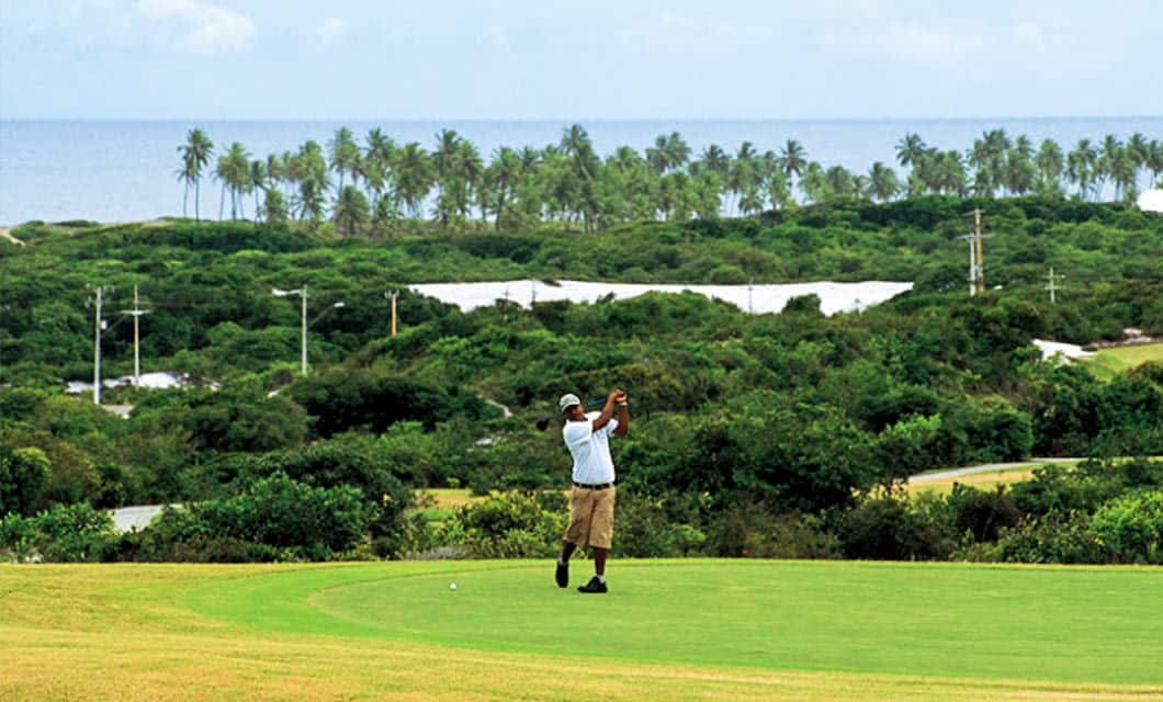 A golfer in a white shirt and khaki shorts swings a club on lush green golf courses, with dense greenery, palm trees, and the ocean visible in the background under a partly cloudy sky.