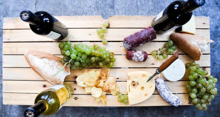 A wooden table with Mendoza wine bottles, green grapes, bread, cheese, salami, potato chips, and a cheese knife arranged for a picnic or wine tasting snack.