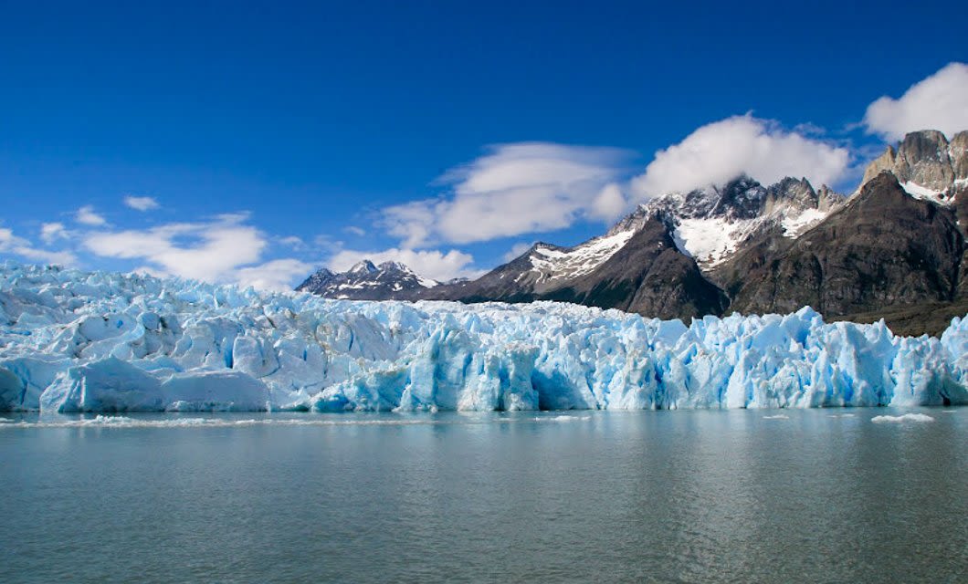 A large glacier with jagged blue ice stretches across the foreground, meeting calm water—an iconic scene along the W Circuit in Torres del Paine. Snow-capped mountains rise beneath a bright blue sky with scattered clouds.