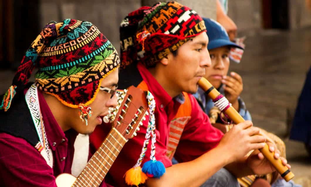 Several musicians wearing colorful, traditional Andean clothing and hats perform on guitar and wooden flute during a vibrant Coast to Coast Tour, all while seated outdoors.
