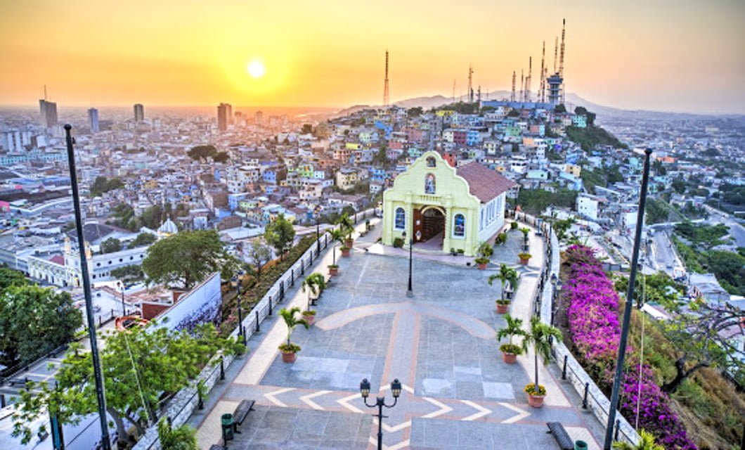 A scenic view of Guayaquil at sunset, featuring the colorful Chapel of Santa Ana on a hilltop—a must-see on any Ecuador tour—overlooking the city with blooming flowers, palm trees, and tall buildings in the background.