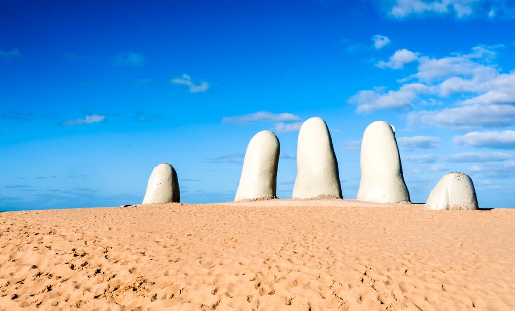 Large white sculpted fingers rise from sandy ground under a blue sky with scattered clouds, resembling a giant hand partially buried in the desert near Punta del Este, Uruguay.