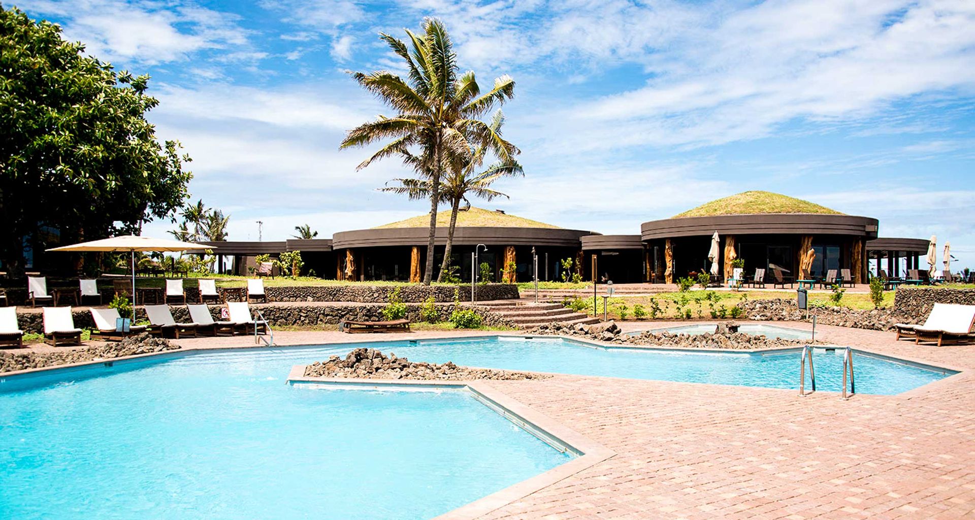 A sustainable hotel, Hangaroa Eco Village Easter Island, features a large outdoor pool, sun loungers, palm trees, and a unique circular building with a grass-covered roof under a blue sky with scattered clouds.