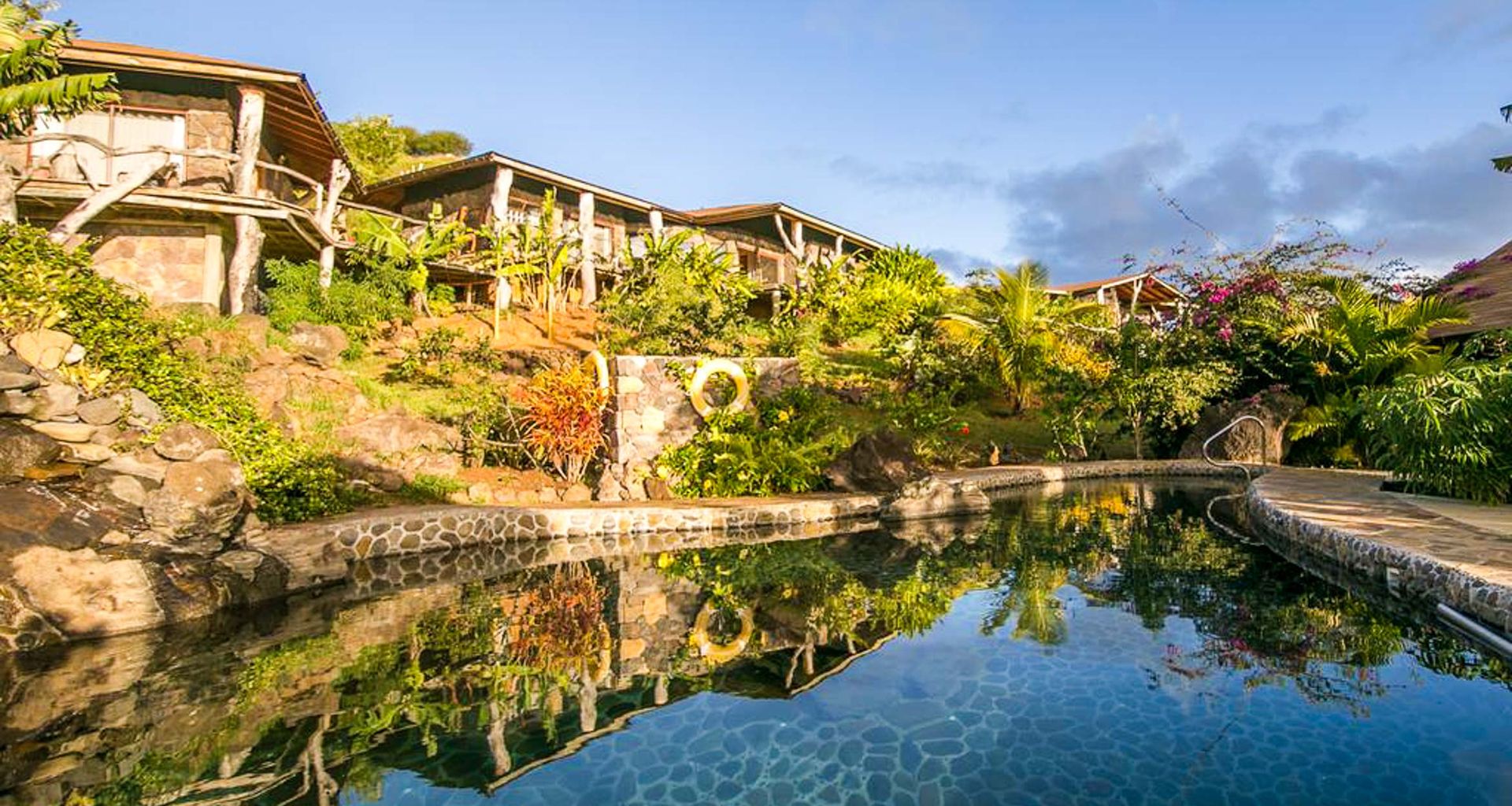 A serene outdoor swimming pool with natural stone edges reflects tropical plants and several rustic Hare Noi wooden lodges set on a hillside under a blue Rapa Nui sky with scattered clouds.