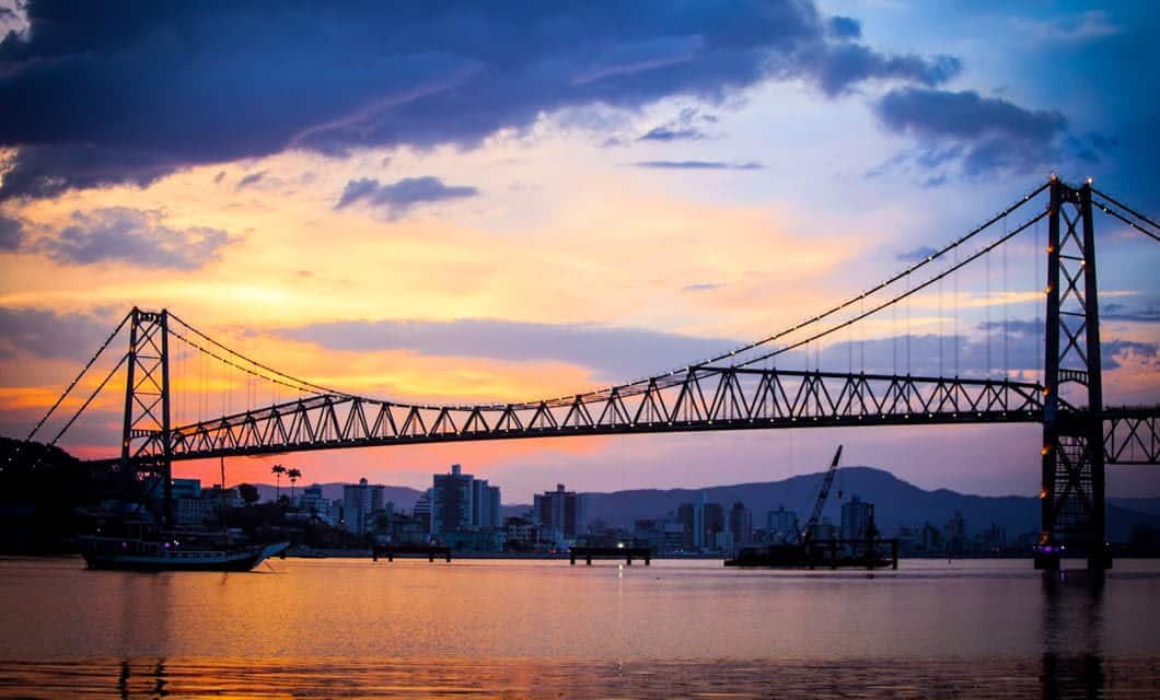 Suspension bridge over calm water at sunset in Florianopolis, Santa Catarina, with a colorful sky of orange, purple, and blue hues. City buildings and distant hills form the backdrop as a boat passes by—perfect for an adventure tour.