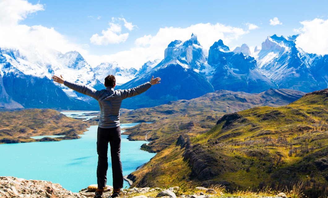 A person stands with arms outstretched overlooking a turquoise lake and dramatic snow-capped mountains in Torres del Paine under a bright sky, surrounded by green hills and rugged landscape—a perfect scene from an unforgettable tour.