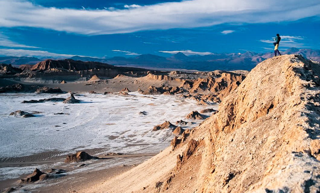 A person stands on a rocky hill overlooking the highlights of Chile’s vast desert landscape, with rugged formations, white salt flats, and distant mountains beneath a blue sky with wispy clouds.