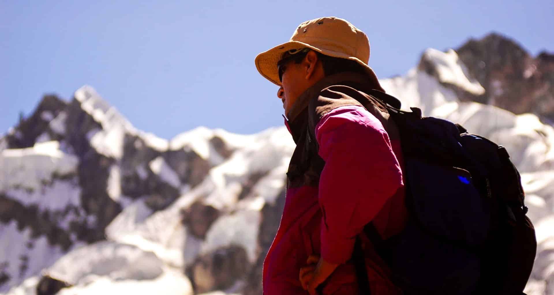 A person wearing a hat, sunglasses, and a backpack stands in front of snow-covered mountain peaks on the Salkantay Trek, gazing into the distance under a clear blue sky on their journey to Machu Picchu.