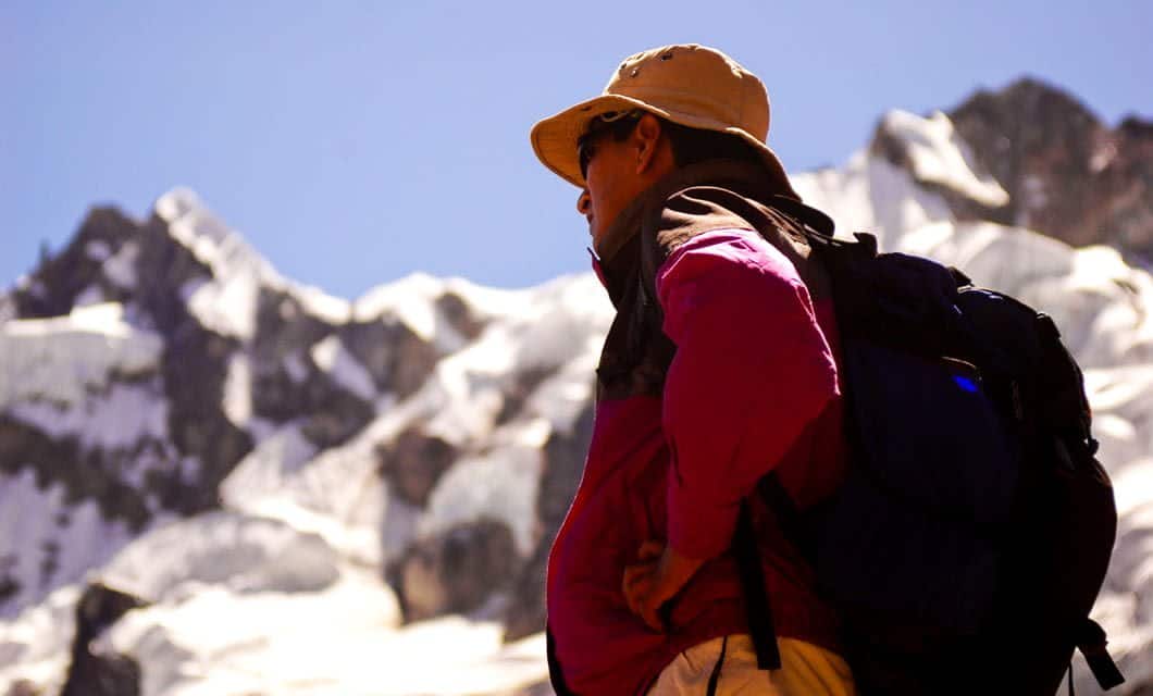 A person wearing a hat, sunglasses, and backpack stands in front of snow-covered mountain peaks under a clear blue sky, looking up toward the mountains along the scenic Salkantay Trek near Machu Picchu.