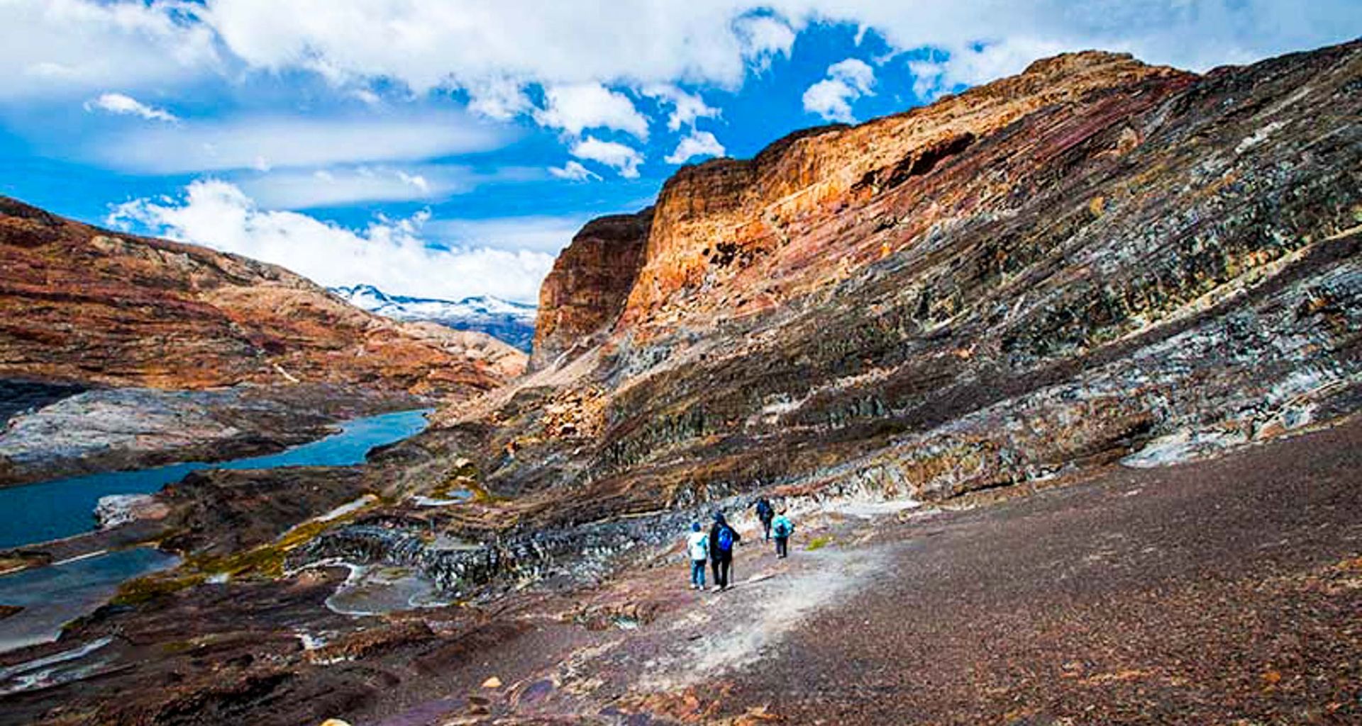 Hikers walk along a rocky mountain path in Patagonia, with colorful, layered cliffs and a river or lake below under a bright blue sky with clouds near Estancia Cristina, Argentina.
