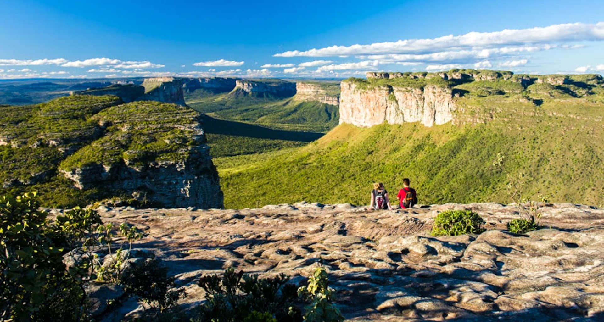 Two people sit on a rocky outcrop in Chapada Diamantina, overlooking lush green valleys and dramatic cliffs under a bright blue sky—an inviting scene for hiking amid breathtaking landscapes.