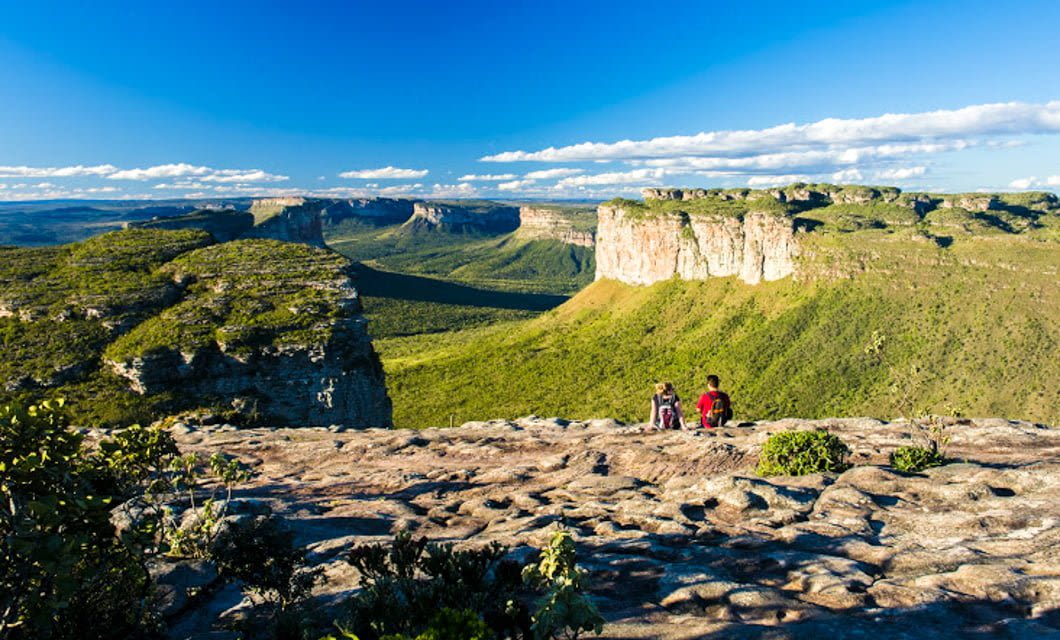 Two people rest on a rocky cliff in Chapada Diamantina, overlooking a vast green valley and dramatic flat-topped mountains under a bright blue sky—a perfect hiking escape sparkling with the allure of diamonds.